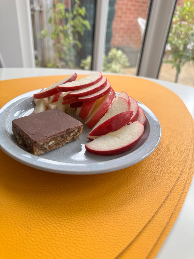 A gray plate on a yellow table with sliced red apples and a chocolate brownie, with windows and outdoor plants visible in the background